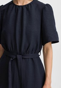 Abito blu navy realizzato in tessuto testurizzato, con maniche a sbuffo corte, scollatura arricciata e cintura in vita. Presenta una vestibilità comoda e una silhouette fluida.