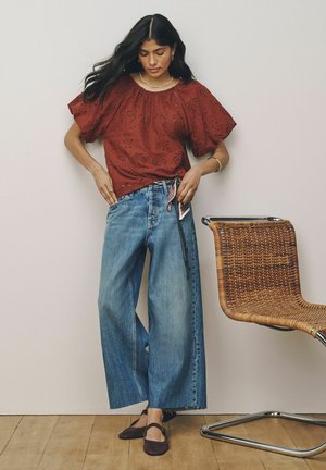 Frau richtet blaue Jeans mit weitem Bein und einem gebundenen Tuchgürtel, trägt eine rostfarbene Lochbluse und schwarze Ballerinas, steht neben einem geflochtenen Stuhl.