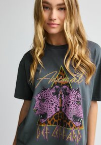 Graues, übergroßes T-Shirt mit einem grafischen Druck von lila Leoparden auf einem Leopardenmuster-Hintergrund, mit den Texten "Animal" und "Def Leppard".