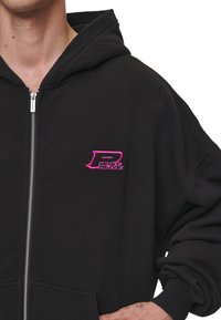 Čierna mikina s kapucňou vyrobená z mäkkej tkaniny, s strieborným zipsom, prednými vreckami a malým ružovým vyšívaným logom na hrudi.