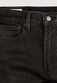 Gros plan d'un jean en denim noir montrant la poche avant, la boucle de ceinture et la fermeture par bouton métallique.