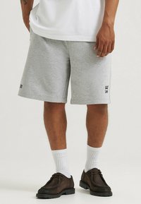 Pantaloni corti grigi in cotone con una vita elastica, tasche laterali e un piccolo logo nero vicino all'orlo, abbinati a calze bianche e scarpe scure.