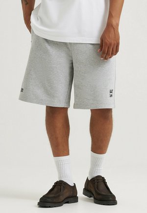 Pantaloni corti grigi in cotone con una vita elastica, tasche laterali e un piccolo logo nero vicino all'orlo, abbinati a calze bianche e scarpe scure.