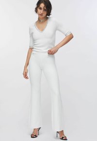 Donna con capelli corti e ricci indossa una maglia bianca a scollo a V con rifiniture argento, pantaloni bianchi a zampa ad alta vita e sandali neri open-toe con dettagli dorati.