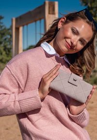 Mladá žena v růžovém svetru drží světle šedou prošívanou peněženku venku, v pozadí se nachází dřevěná houpačka.