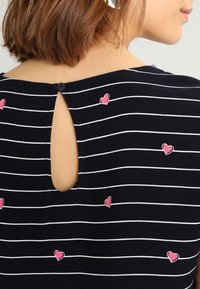 Femme portant un haut noir à rayures horizontales blanches et de petits motifs en forme de cœur roses, avec un décolleté en forme de goutte d'eau fermé par un bouton.