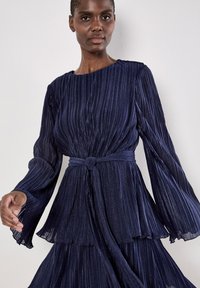Granatowa sukienka plisowana z długimi dzwonowymi rękawami, związaną w talii oraz warstwową spódnicą. Teksturowany materiał z delikatnym połyskiem i węzłem jako akcentem.
