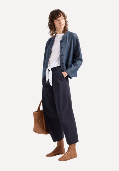 Blauwe gestructureerde jas, witte geknoopte blouse, donkere broek, bruine geperforeerde tas en bijpassende enkellaarsjes vormen een casual outfit.