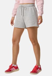 Ben iført lysegrå shorts med snøre, røde Adidas-sneakers med hvide striber, hvide sokker og en pink cropped hoodie.