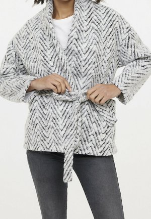 Osoba wiążąca pasek na szarej i białej teksturowanej, wzorzystej dzianinowej kardiganie noszonej na białym topie i ciemnoszarych spodniach.