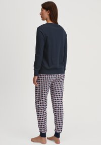 Vrouw staat blootsvoets zijwaarts gewend, gekleed in een lange mouw marineblauw shirt en geruite pyjamabroek met rode en witte accenten.