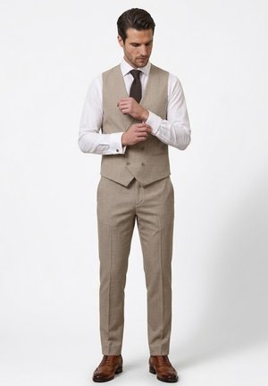 Hombre ajustando el gemelo, vestido con chaleco de traje beige, pantalones a juego, camisa blanca de vestir, corbata oscura y zapatos de vestir marrones sobre fondo blanco.