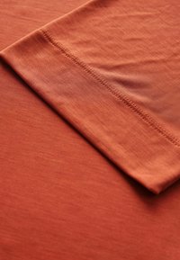 Roestkleurige stof met een gladde textuur, voorzien van een omgevouwen zoom en zichtbare stiksel-details. Het materiaal lijkt lichtgewicht en zacht.