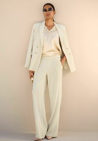 Vrouw in een crèmekleurig pak met wijde broekspijpen, satijnen top met kant, zonnebril en huidskleurige hakschoenen, staand tegen een beige muur.