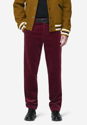 Person trägt burgunderfarbene Cordhosen, schwarzen Gürtel, schwarze Schuhe, schwarzes Hemd und eine braune Collegejacke mit gestreiften Bündchen und Taschen.
