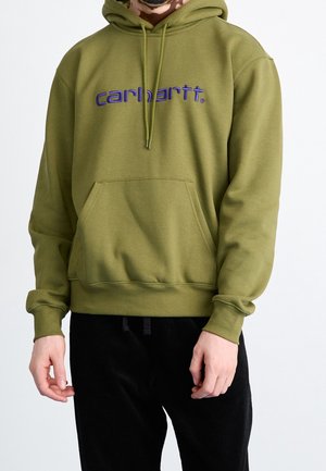 Mężczyzna ubrany w oliwkowo-zieloną bluzę z kapturem z niebieskim logo „Carhartt” na środku klatki piersiowej i czarne spodnie, stojący na jednolitym białym tle.