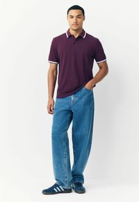 Polo di colore bordeaux con rifiniture bianche, abbinato a jeans larghi in denim blu. Il modello si trova in posizione casuale, con le mani in tasca, indossando sneakers blu.