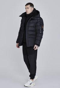 Sort pufferjakke med høj krave, lynlås lukning og logo patch på ærmet. Sammen med sorte sweatpants og hvide sneakers.
