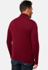 Ein Mann trägt einen langärmeligen, gerippten burgunderfarbenen Pullover mit hohem Kragen und dunkelblaue Jeans, während er von einem einfarbigen Hintergrund abgewandt steht.