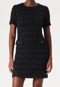 Kortärmad svart tweedklänning med ett strukturerat mönster, som har två framfickor och små metalliska detaljer.