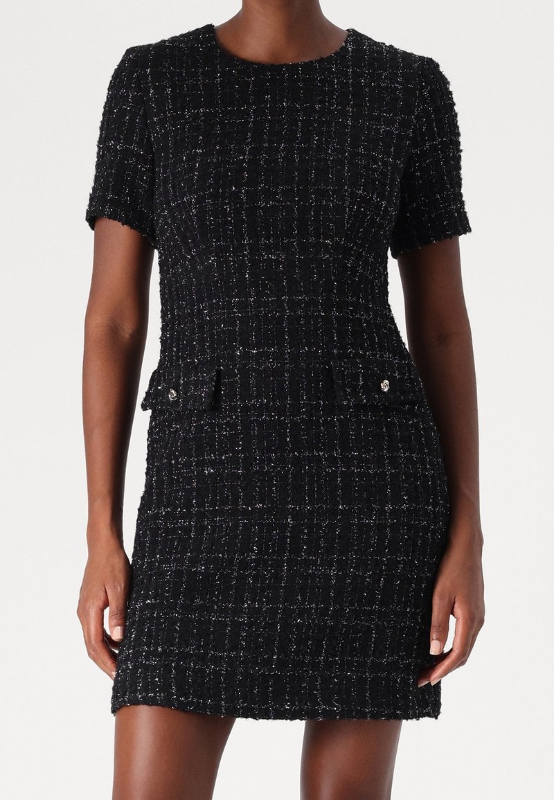 Kortärmad svart tweedklänning med ett strukturerat mönster, som har två framfickor och små metalliska detaljer.