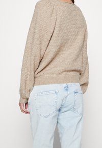 Eine Person trägt einen lockeren beige Strickpullover und leichte blaue Jeans, von hinten vor einem schlichten Hintergrund gezeigt.