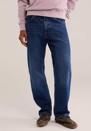 Persona che indossa jeans blu scuro a gamba dritta, sneakers marroni con lacci e una felpa rosa chiaro, in piedi su uno sfondo neutro.