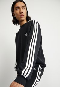 Svart sweatshirt i mjukt material med tre vita ränder på ärmarna, rund halsringning och liten logga på bröstet och benet.