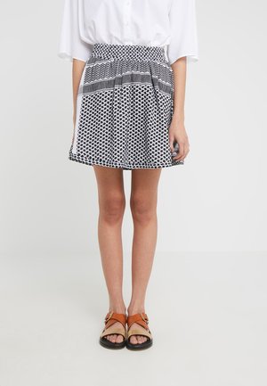 Kobieta ubrana w krótką spódnicę w czarno-biały wzór, białą bluzkę i brązowe sandały z odkrytymi palcami, stojąca na tle jednolitego jasnego tła.