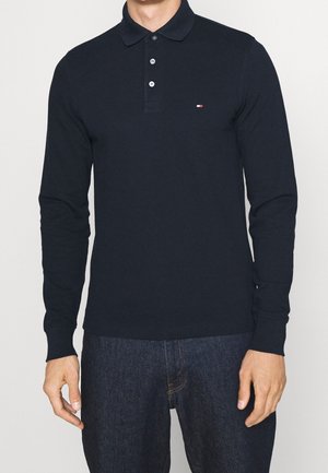 Man draagt een nauwsluitende zwarte polo met lange mouwen, drie knopen en een klein logo op de borst, gecombineerd met een donkere spijkerbroek.