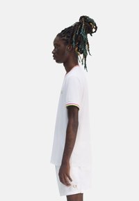 Camiseta blanca de manga corta con acentos de rayas rojas, verdes y amarillas en el cuello y las mangas, combinada con pantalones cortos blancos, que presentan un detalle de logo.