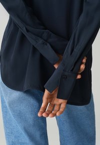 Camicia blu navy con bottoni, maniche lunghe e orlo curvo, abbinata a pantaloni in denim azzurro chiaro. Tessuto morbido e polsini aderenti sono ben visibili.