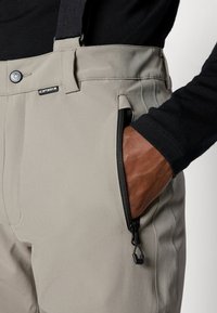 Pantaloni bej, cu talie înaltă, prevăzuți cu bretele negre ajustabile, având un buzunar lateral cu fermoar și un finisaj de țesătură netedă și durabilă.