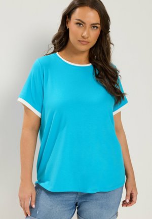 Frau mit langen dunklen Haaren, die ein locker sitzendes hellblaues T-Shirt mit weißer Umrandung und hellblaue Jeans vor einem schlichten Hintergrund trägt.
