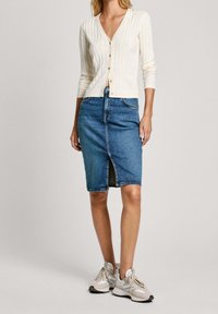 Cardigan bianco a coste con scollo a V, abbinato a una gonna a tubino in denim blu con spacco frontale. Modella che indossa sneaker chiare.