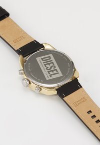 Reloj de acero inoxidable dorado con correa de cuero genuino negro. Presenta tres botones plateados y una tapa de cristal que muestra el logo "DIESEL".
