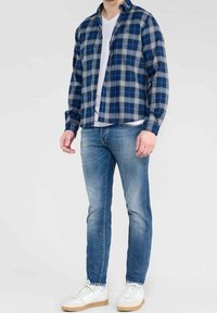 Mies, joka on pukeutunut sinivalkoiseen ruudulliseen paitaan, valkoiseen t-paitaan, kapeisiin sinisiin farkkuihin ja valkoisiin lenkkareihin, joissa on vaaleanruskeat pohjat, seisoo yksinkertaista taustaa vasten.