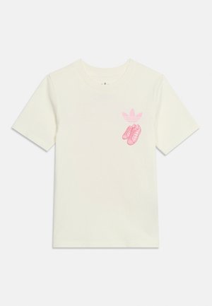 Balta īsroku T-krekls ar nelielu rozā Adidas logotipu un rozā sporta apavu grafiku kreisajā krūtīs.