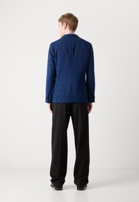 Modra karirana blazer s prilagojenim krojem, v kombinaciji z črnimi hlačami širokega kroja. Blazer ima obrobljen ovratnik in tri gumbe.