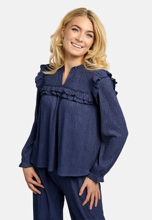 Smehljiva ženska z blond lasmi nosi teksturirano temno modro bluzo z volančki in ujemajoče se hlače, pozira na enostavni svetli podlagi.