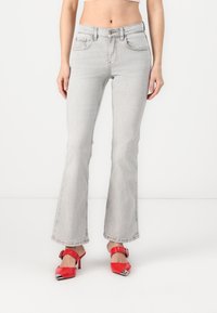 Jean gris évasé avec une texture légèrement délavée, comportant cinq poches et une fermeture à bouton, assorti à des chaussures à talons rouges avec des accents en argent.