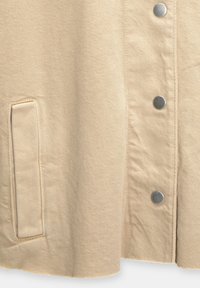 Beige jas gemaakt van textuurstof, voorzien van zilveren drukkers, zijzak en een gladde zoom.