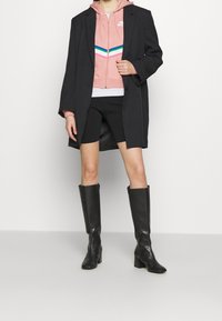 Svart överdimensionerad kavaj över en ljusrosa zip-hoodie med färgglada randiga detaljer, svarta cykelshorts och knähöga svarta klackboots.