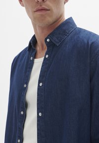 Camisa de mezclilla en azul oscuro, con cuello abotonado y botones plateados. Llevada sobre un top de tirantes de canalé blanco. Tejido texturizado.