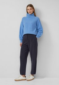 Blauwe textuursweater met hoge col, donkere op maat gemaakte broek en witte sneakers met beige accenten. Eenvoudige, moderne silhouet.