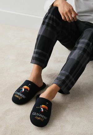 Osoba nosící černé a šedé kostkované kalhoty sedí na koberci, nohy má obuté v černých pantoflích Guinness s barevným logem toucana.