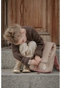 Beige schoudertas met roze accenten, gemaakt van textuurstof. Meisje in een bruine trui en crèmekleurige broek past de tas aan terwijl ze op stenen trappen zit.