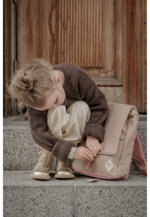 Beige schoudertas met roze accenten, gemaakt van textuurstof. Meisje in een bruine trui en crèmekleurige broek past de tas aan terwijl ze op stenen trappen zit.