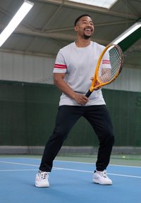 Grå t-skjorte med røde striper på ermene, svarte treningsbukser og hvite tennissko, holder et gult tennistrå på en blå bane.