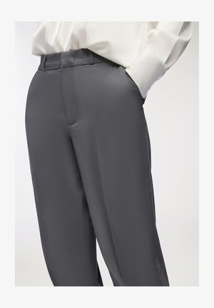 Pantaloni in satin grigio con una texture liscia, caratterizzati da una vestibilità sartoriale e tasche laterali, abbinati a una camicia bianca con bottoni.